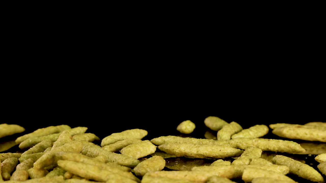 Puffed green pea chips fall and pile up in slow motion against a black background