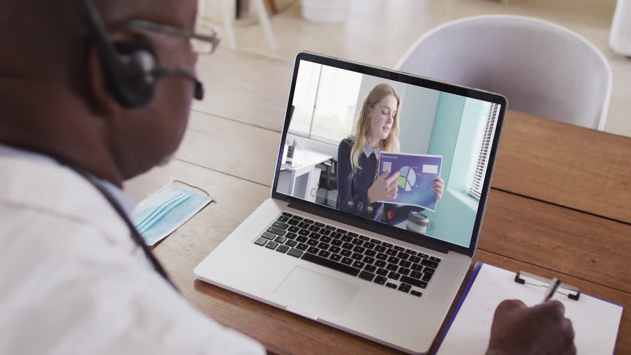 Afroamerikansk mandlig læge med headset på, der tager noter under et videoopkald på sin bærbare computer