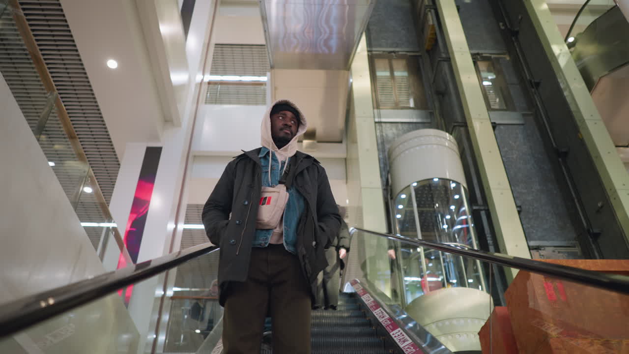 Young man in hoodie, hands in pockets, descends escalator in modern shopping mall. Escalator lined with red caution tape, man carries small shoulder bag. Urban, contemporary, casual atmosphere