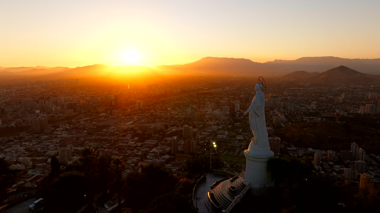 Forward-moving drone at sunset flies past the Sanctuary on San Cristóbal Hill, revealing the giant Immaculate Conception statue above Santiago and the Andes in warm golden haze