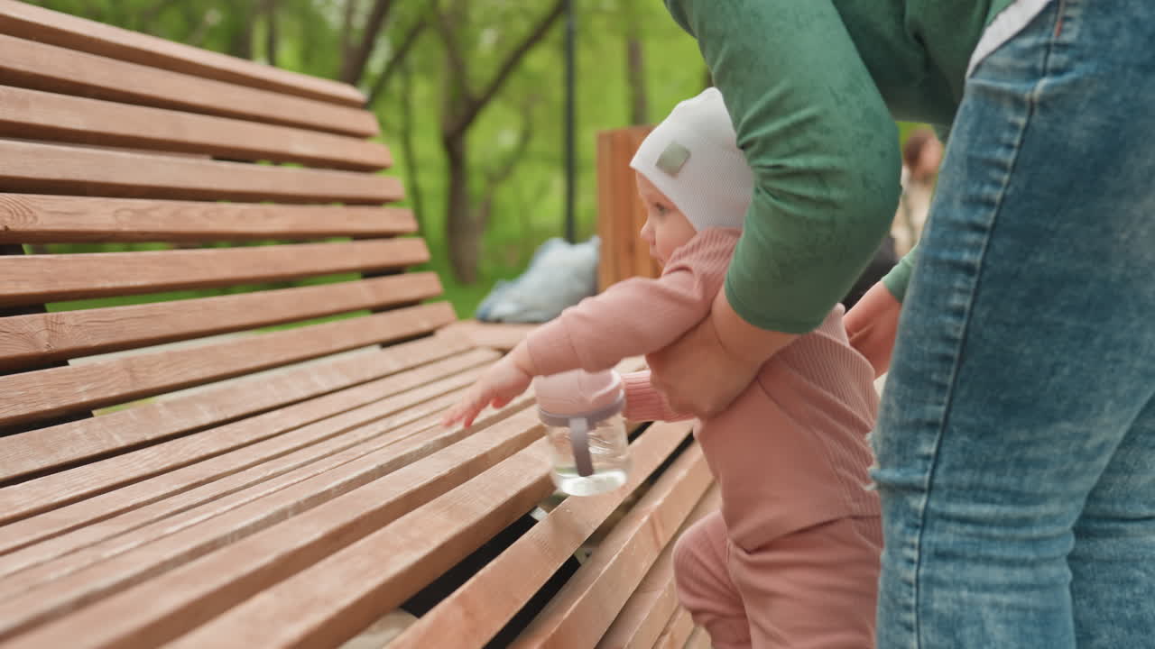 Father Aids Baby, Supporting Child Near Park Bench, Father Assists Infant With Support By Park Bench During Outing, Nurturing Moment As Father Helps Baby Stand Beside Wooden Park Bench