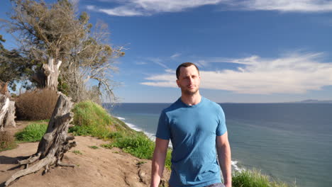 A-man-walking-on-the-edge-of-an-ocean-cliff-on-a-nature-hiking-trail-with-an-epic-beach-view-on-a-sunny-day-in-Santa-Barbara,-California-SLOW-MOTION