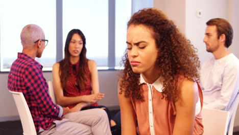 Upset-woman-sitting-on-chair-with-colleagues-discussing-in-background