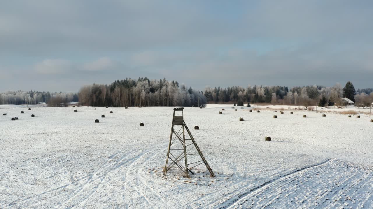 Wildlife animal hunting tower in the middle of field on a sunny winter day. Snow covered ground with hay rolls and frozen forest in the background.