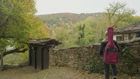 Female-musician-stands-admires-village-Autumn-forest-scenery-pull-shot