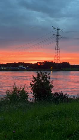 vertical timelapse of stunning sunset over a river in the netherlands as golden sky and orange clouds drift above a transmission tower silhouetted against the fading light.