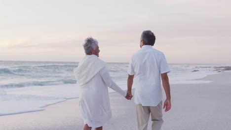 Back-view-of-happy-hispanic-just-married-senior-couple-walking-on-beach-at-sunset