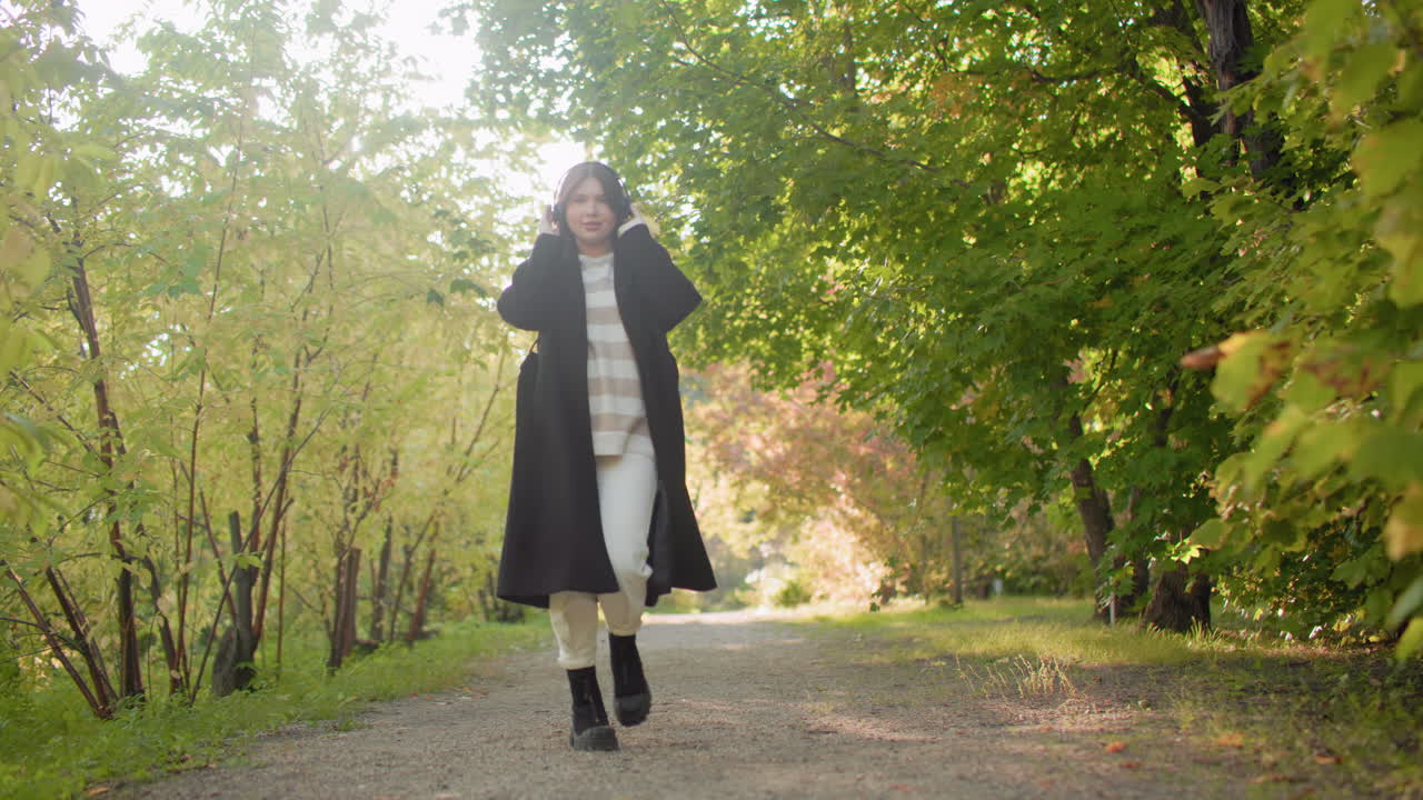 Playful lady enjoying walk with headset on, spinning round in rhythmic expression along scenic autumn pathway, surrounded by sunlight filtering through lush green trees and golden seasonal foliage