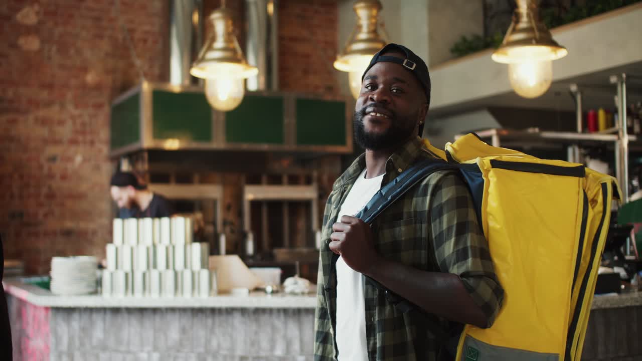 A food delivery man, a person with Black skin color, in a plaid shirt, poses and smiles in a doner market. Video filmed in high quality