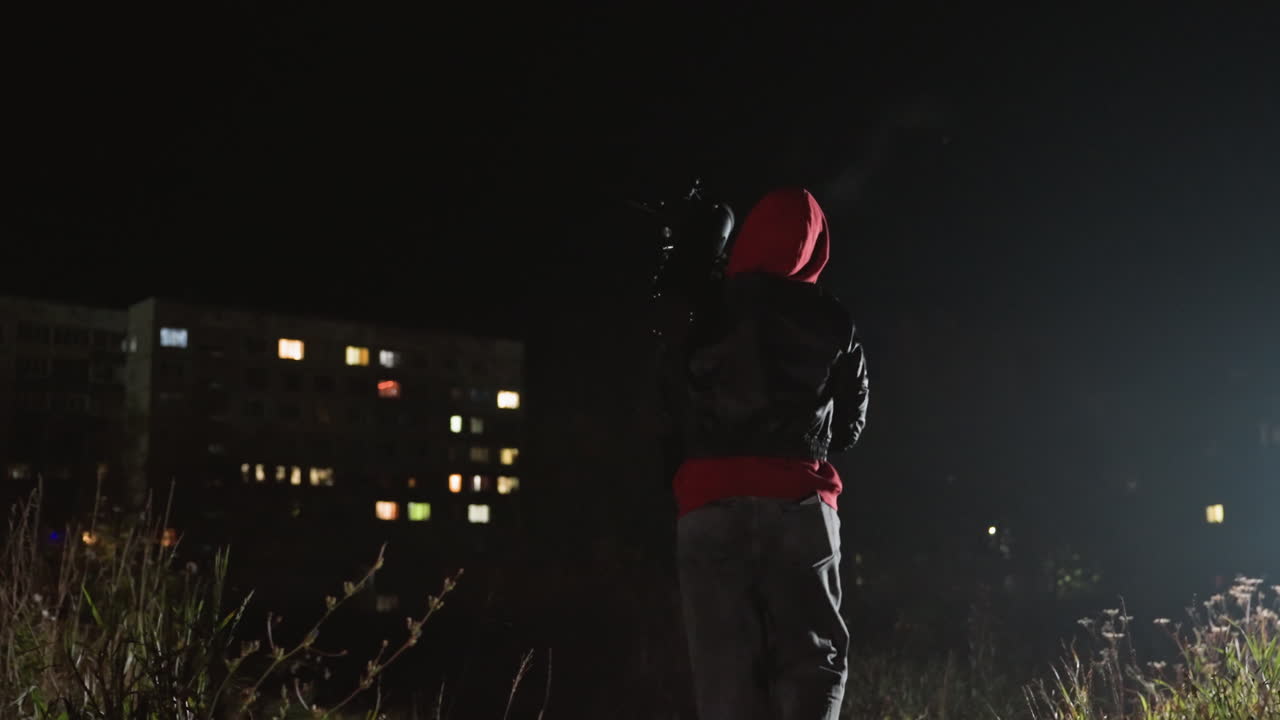 Una persona con un telescopio di notte monta un treppiede in un terreno incolto vicino a dei condomini; una figura incappucciata regola l'oculare sotto la luce della città; un'illuminazione cinematografica intensa evoca una tranquilla curiosità e spirito scientifico