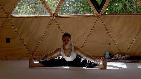 Latina woman practicing yoga in a geometric studio, focusing on mindfulness and calm breathing sitting down with legs stretched and arms to feet