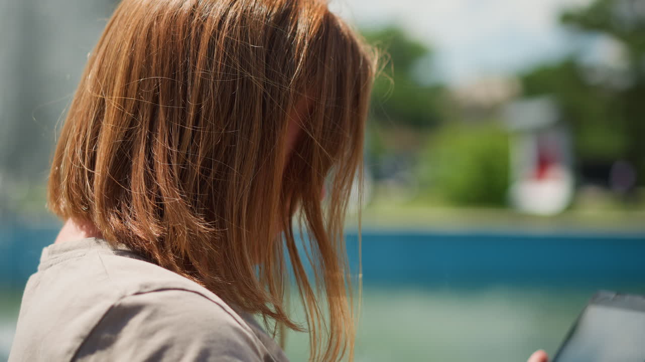 Young lady admiring something on phone with smile under bright sunlight near fountain, soft breeze moving her hair, peaceful mood expressing curiosity surrounded by natural green summer background