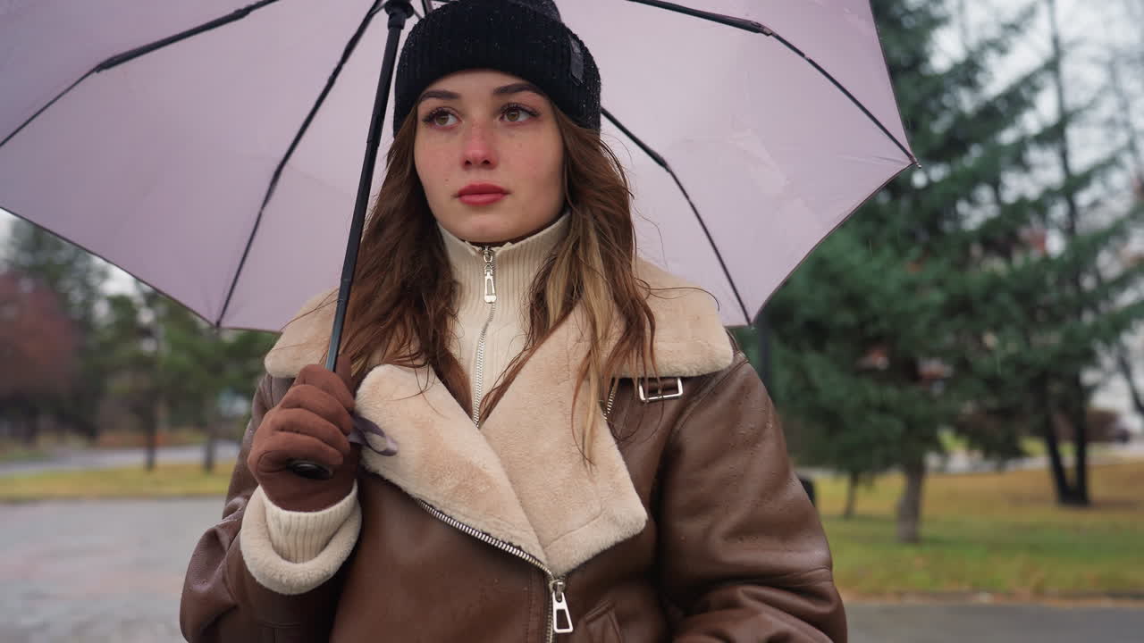 Student walking in park on cold overcast day, holding umbrella, dressed in black knit cap and brown shearling jacket, surrounded by evergreen trees, with peaceful atmosphere and soft winter lighting