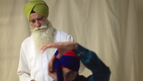 studio-aufnahme eines älteren sikh-mannes, der einem jüngeren sikh hilft, stoff für einen turban gegen einen einfachen hintergrund zu binden. aufnahme in echtzeit 2