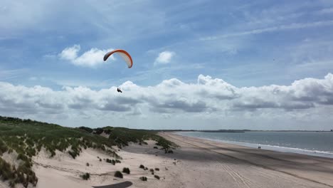 drone backing away from paraglider drifting above lush seafront sand dunes