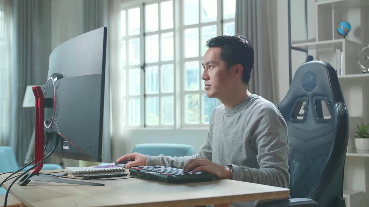 Asian Man In Long Sleeved T-Shirt Using Desktop Computer On A Table For Working At Home.