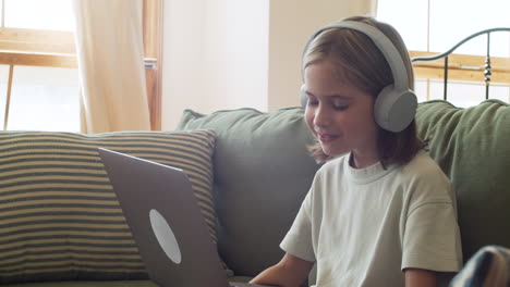 close up of a little blonde girl with headphones having fun while watching at the laptop screen seated on the sofa