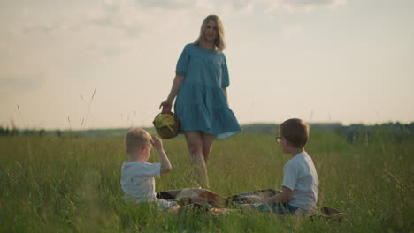 a woman in a flowing blue dress walks toward two young boys, dressed in white tops, who are sitting on a scarf in a grassy field. she carries a basket of fruit, creating a peaceful picnic