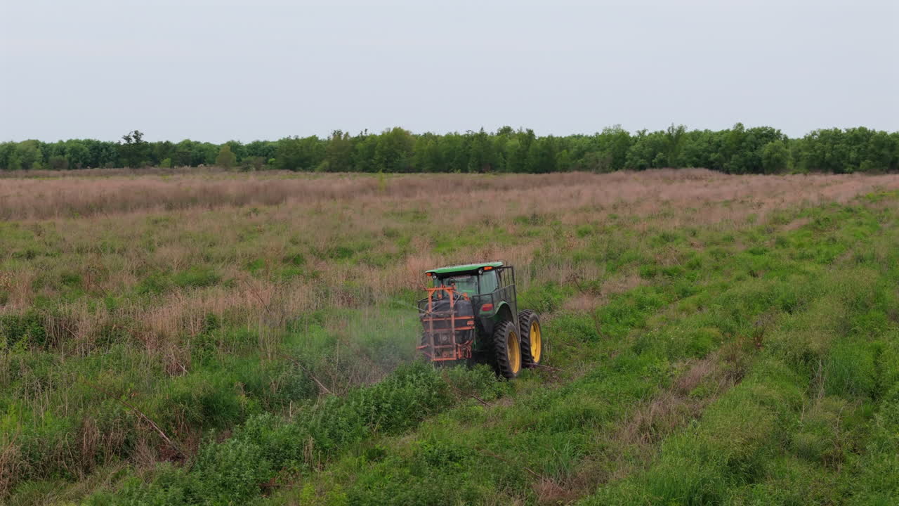Slow drone clip panning over tractor while it spray fertilizes fields. Large extension crops surrounded by trees, with sky full of clouds.