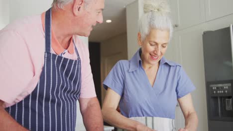 Glückliches-Kaukasisches-älteres-Paar-Mit-Schürzen,-Das-Gemeinsam-In-Der-Küche-Kocht