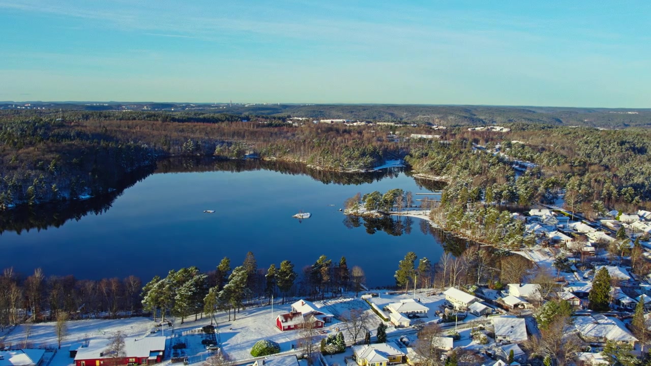 Luftaufnahmen mit Drohne über einer gefrorenen Seenlandschaft, schneebedeckten Häusern, Waldlandschaft und ruhigen Wintergewässern in Öjersjö, Schweden