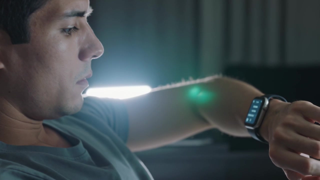 Glancing adult male raising left wrist on sofa at night, inspecting smartwatch tiles