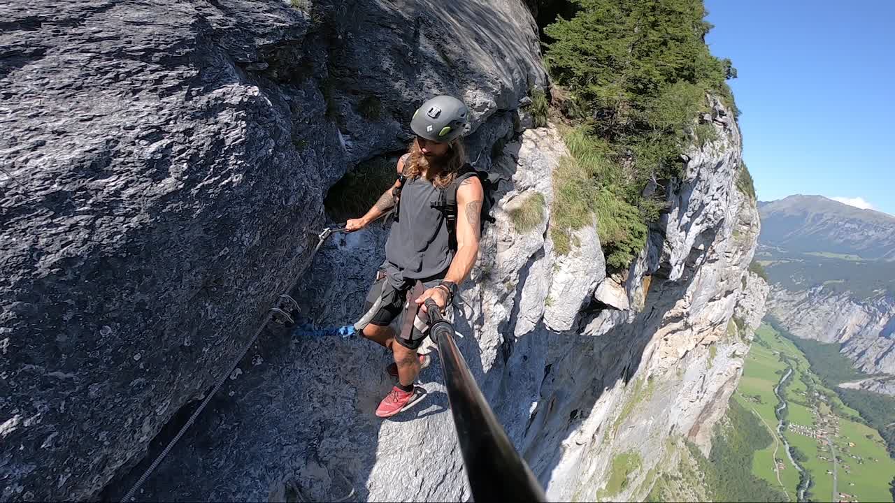 Young bearded man with tattoos is walking down carefully and slowly on the very narrow path on the cliffside of Via Ferrata route in Switzerland. Hot day with harch sunlight and clear blue sky.