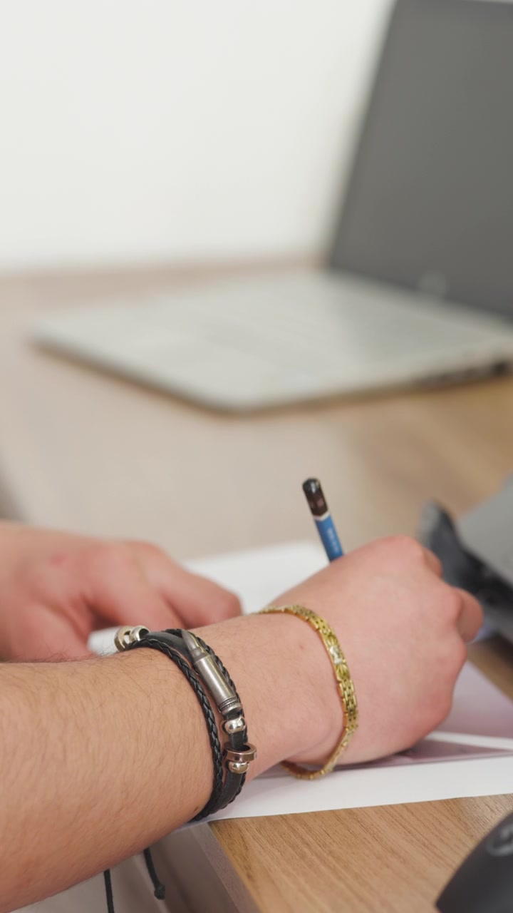 L'inquadratura evidenzia in primo piano una mano con braccialetti in pelle e metallo che tiene una matita blu sopra fogli stampati.