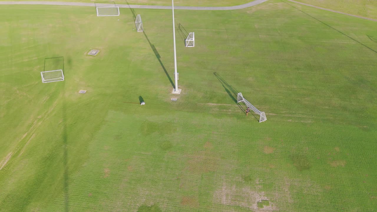 Drone footage captures a soccer player moving across a sunlit field, highlighting the expansive green landscape and goalposts