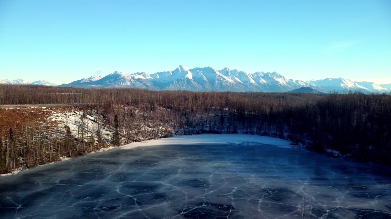 Low altitude drone footage climbing dramatically over trees in the lower left frame, then ascending smoothly across a frozen lake's surface. The camera rises gradually, revealing deep