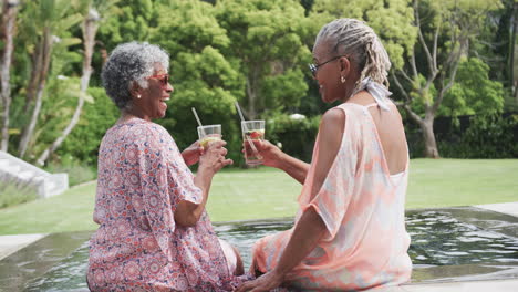 Felices-Amigas-Afroamericanas-Mayores-Haciendo-Un-Brindis-Con-Bebidas-Sentadas-Junto-A-La-Piscina,-Cámara-Lenta