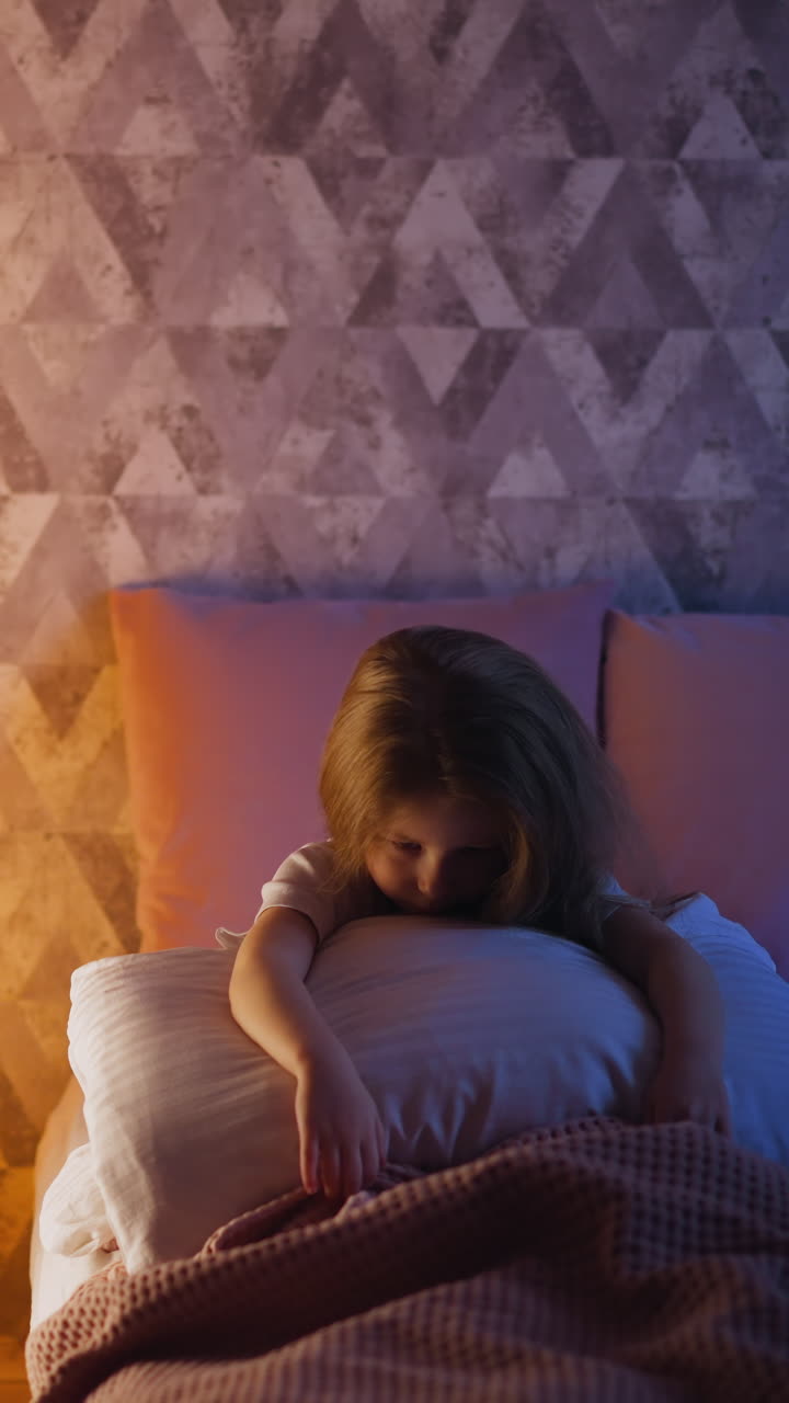 Tired little girl hugs huge pillow sitting on comfortable bed in semidark room illuminated by night lamp glowing inside play tent in evening slow motion