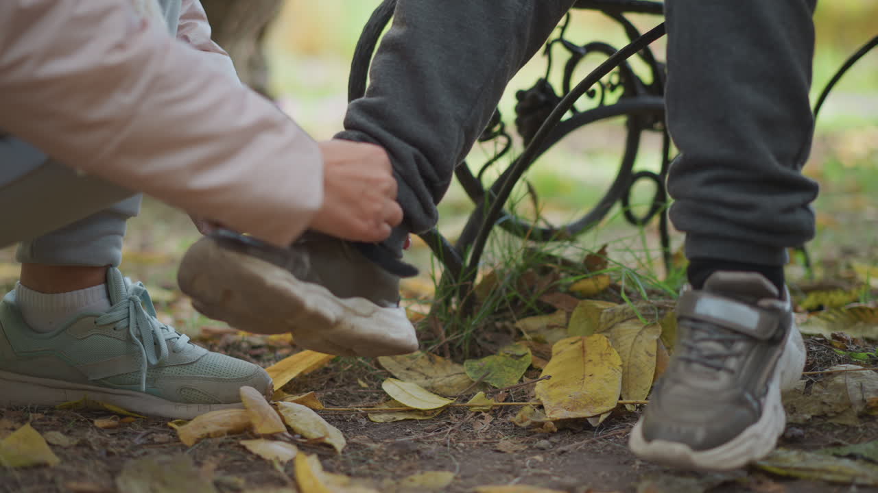 close up of mother gently adjusting kid shoe strap while crouching on fallen autumn leaves near iron bench, showcasing love, attention, and seasonal warmth in outdoor natural park setting