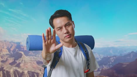 close up of asian male hiker with mountaineering backpack looking at camera and making stop hand sign while traveling at the top of mountain