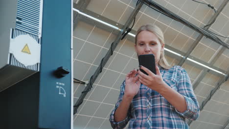 the woman uses a smartphone stands at the inverter under the panels of the house