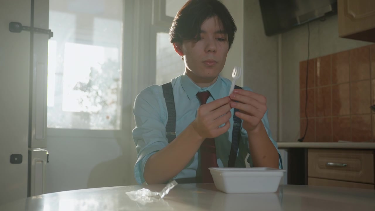 日当たりの良いキッチンでインスタントラーメンを作る、ネクタイ姿のアジア系会社員。調味料の袋を剥がし、スープをかき混ぜ、香りを確かめる集中した表情。レトロなタイル張りのバックスプラッシュ、木製キャビネット、そして丼。