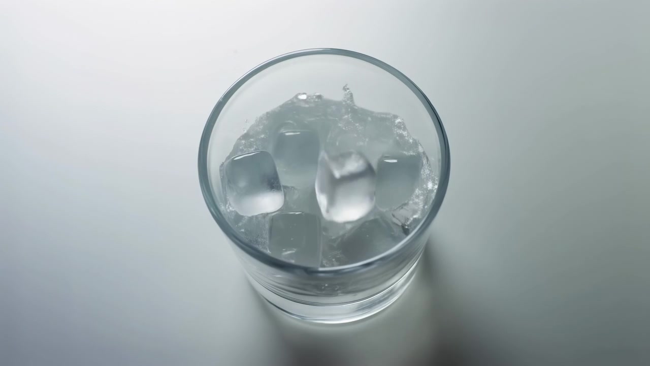 Cubetti di ghiaccio che cadono in un bicchiere d'acqua cilindrico trasparente, formando uno schizzo energico, increspature a corona, goccioline alla deriva e riflessi luminosi su uno sfondo da studio grigio tenue con una vista aerea nitida.