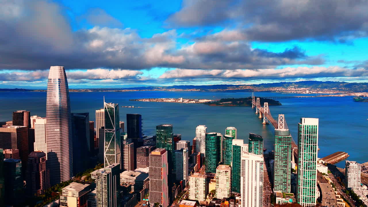 Luftperspektiv af San Franciscos skyline i centrum, med moderne skyskrabere og Bay Bridge i baggrunden under en delvist skyet himmel. Havet er fyldt med lette krusninger.