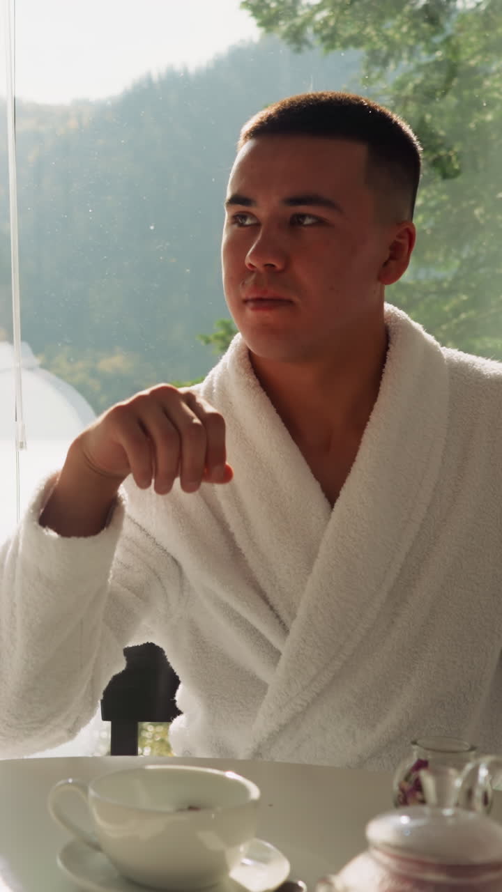 한 남자가 차에 장미 꽃잎을 던지고 컵에 술을 마시며 여인에게 눈을 니다. 테리 바트 로브를 입은 젊은 커플은 아침 식사에서 함께 뜨거운 음료를 마시며 재미있습니다. 편안한 아침 일상