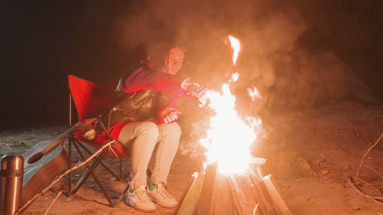 Fogueira em chamas na noite, campista assustado, cadeira dobrável jogada para trás, faíscas e chamas irrompendo perto das pernas, fumaça subindo ao céu, jaqueta vermelha e moletom visíveis, botas na areia, barraca próxima, dramático.