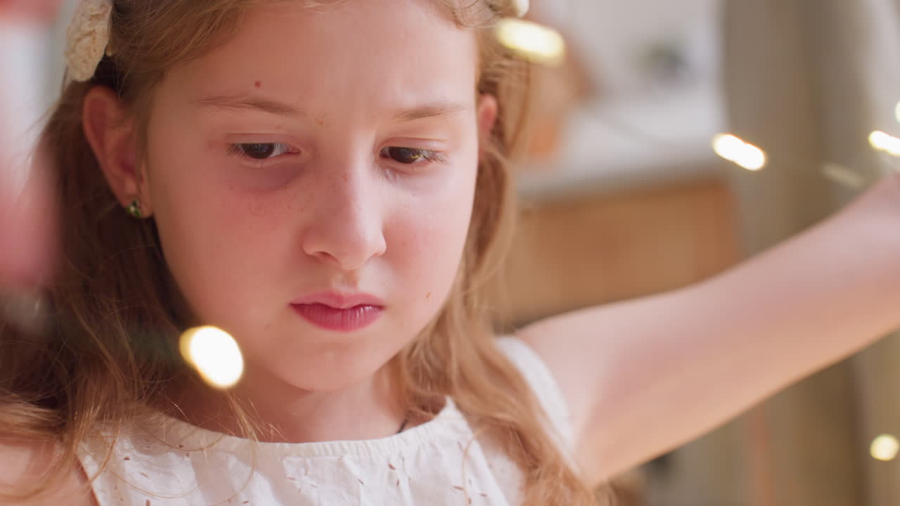Close-up portrait of little girl in white gown focusing on Christmas light while adjusting or fixing it during holiday preparations, looking down with concentration on the task