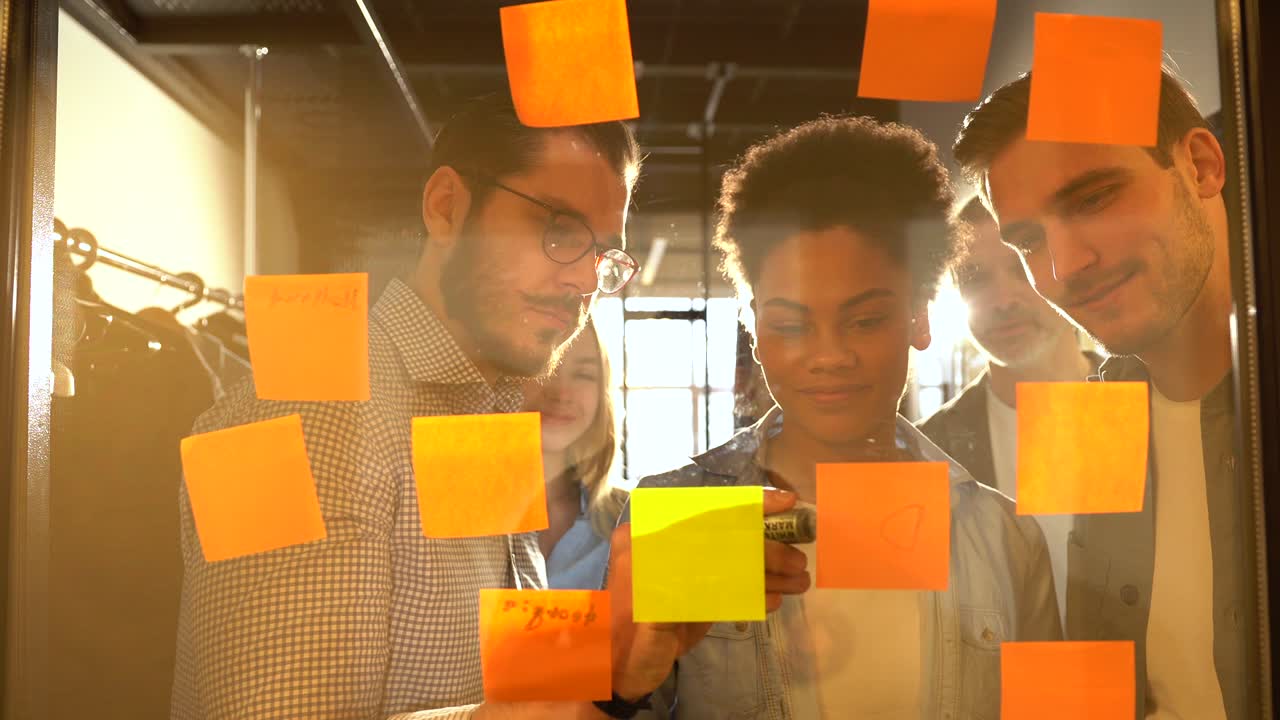 Diverse coworkers group analyzing corporate strategy drawing chart on glass wall with sticky notes. Smiling diverse project managers managing workflow, discussing idea, writing notes on paper stickers