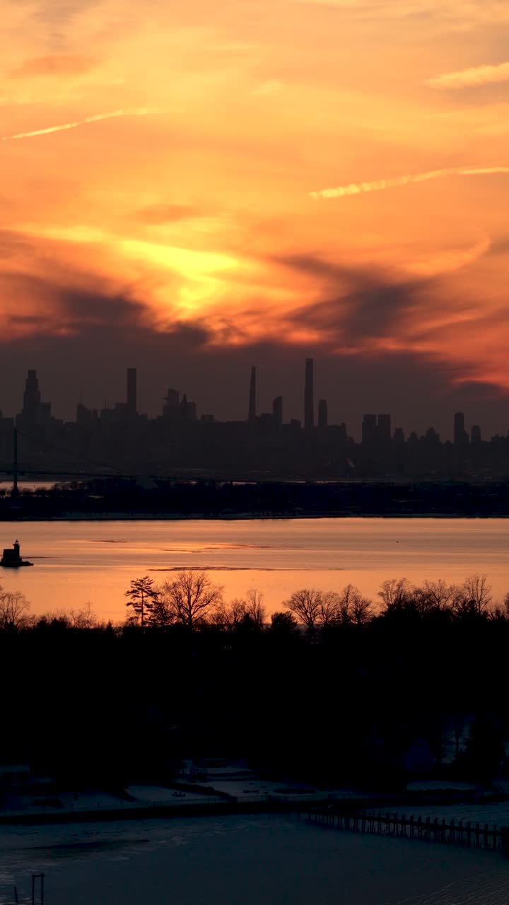 Dramatic golden sunset casting a warm glow over the silhouetted manhattan skyline, with a frozen bay and snow covered ground in the foreground, creating a striking winter urban landscape
