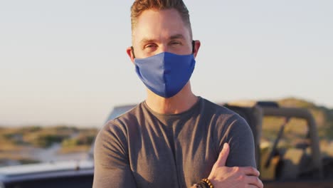 Portrait-of-happy-caucasian-man-in-face-mask-standing-by-car-on-sunny-day-at-the-beach