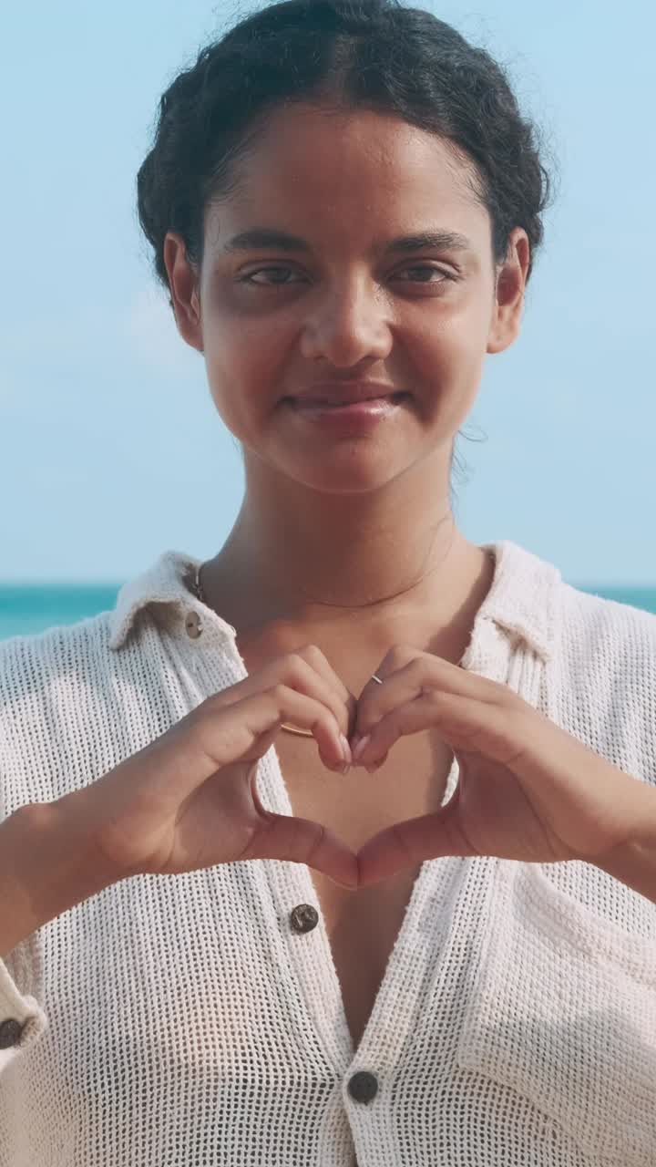 Woman creates heart shape with hands at beach in sunny weather
