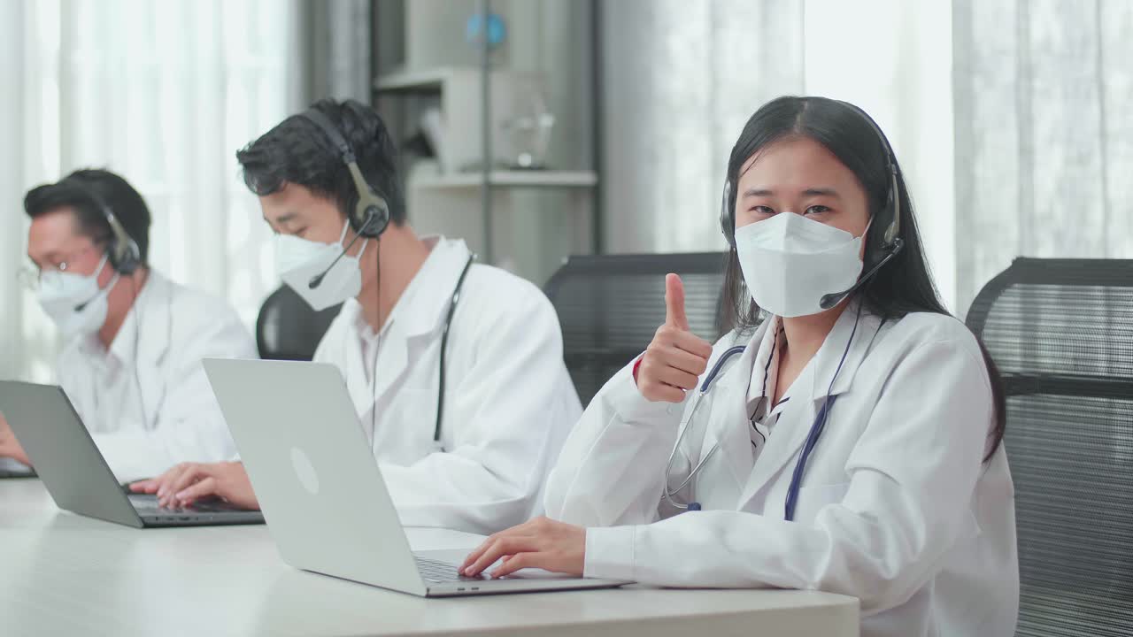 A Woman Of Three Asian Doctors With Stethoscope In Headset And Mask Working As Call Center Agent Look Up From Computer And Thumbs Up While Her Colleagues Are Speaking And Typing During A Call At The Office