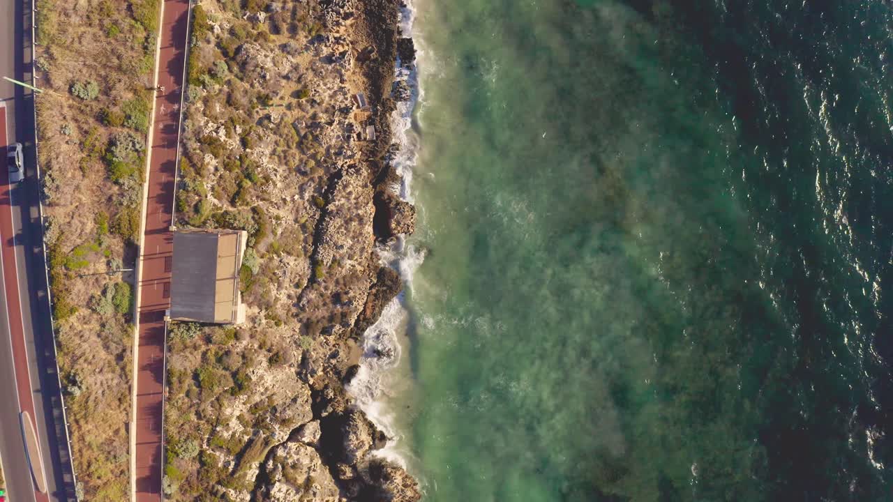 Moving overhead drone top down shot travelling over rocky surf beach and nearby road and bike path