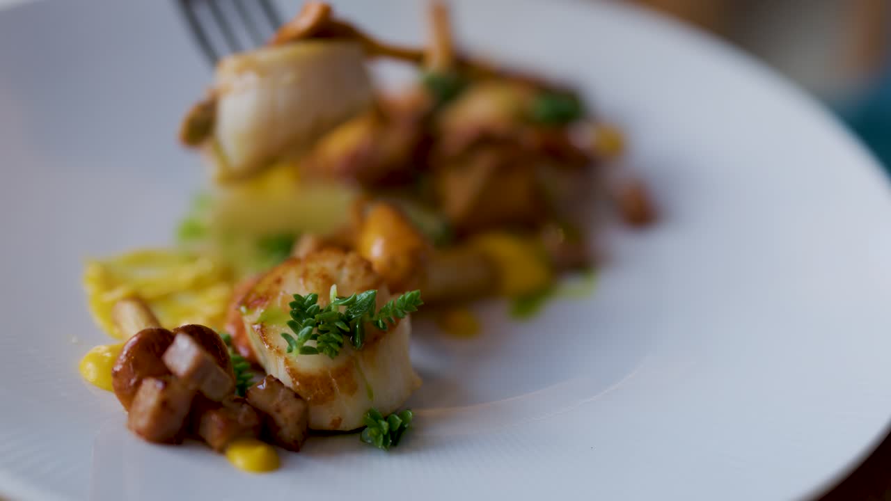 A fork delicately lifts a seared scallop from a gourmet seafood plate, presented on a white dish with elegant garnishes and soft natural lighting