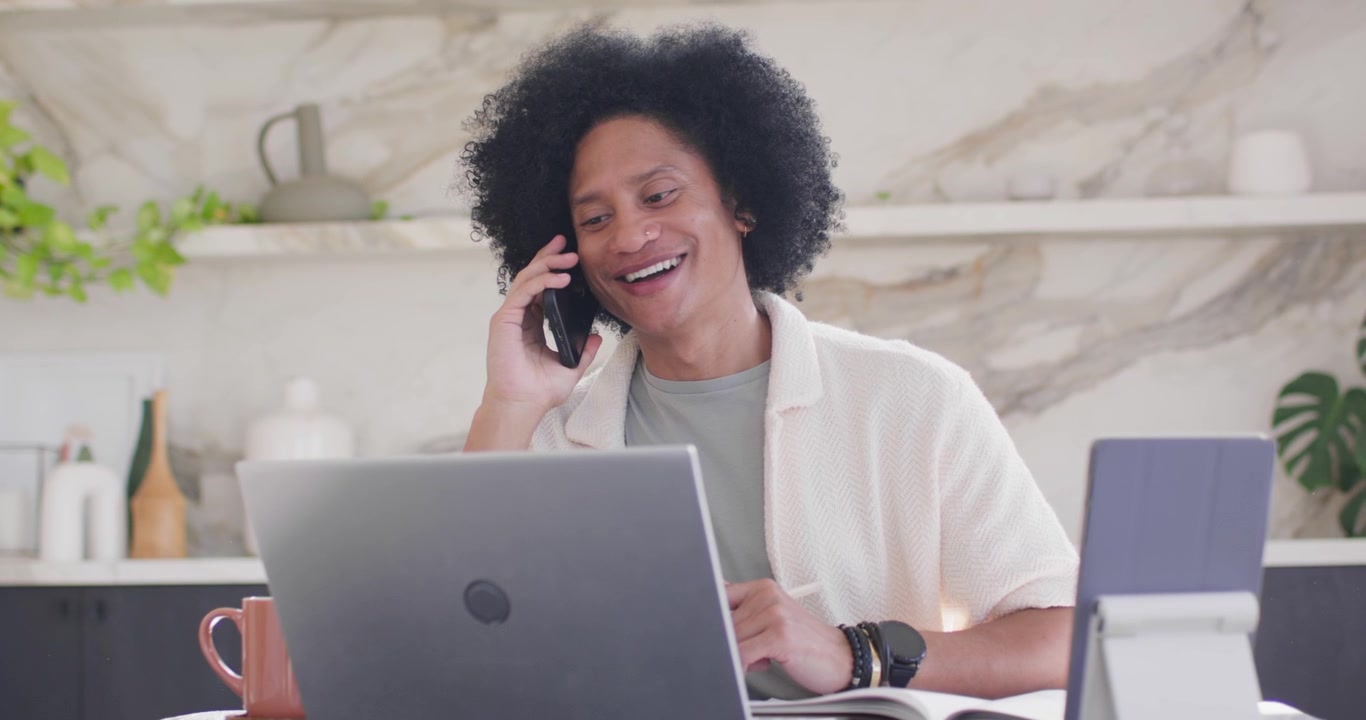 Uomo afroamericano che digita sul laptop in cucina, guardando a destra, prendendo lo smartphone, unendosi a una chiamata