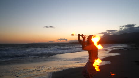 夕暮れのビーチで炎と海の波のスローモーションで燃える火の棒を回転させて踊るシルエットの男